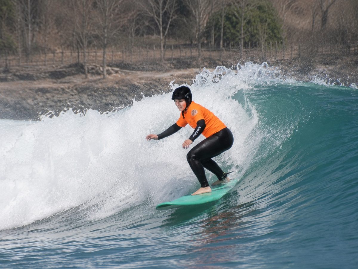 Junge Surferin mit Helm und orangem Shirt auf einer brechenden Welle (Anna Haiss)